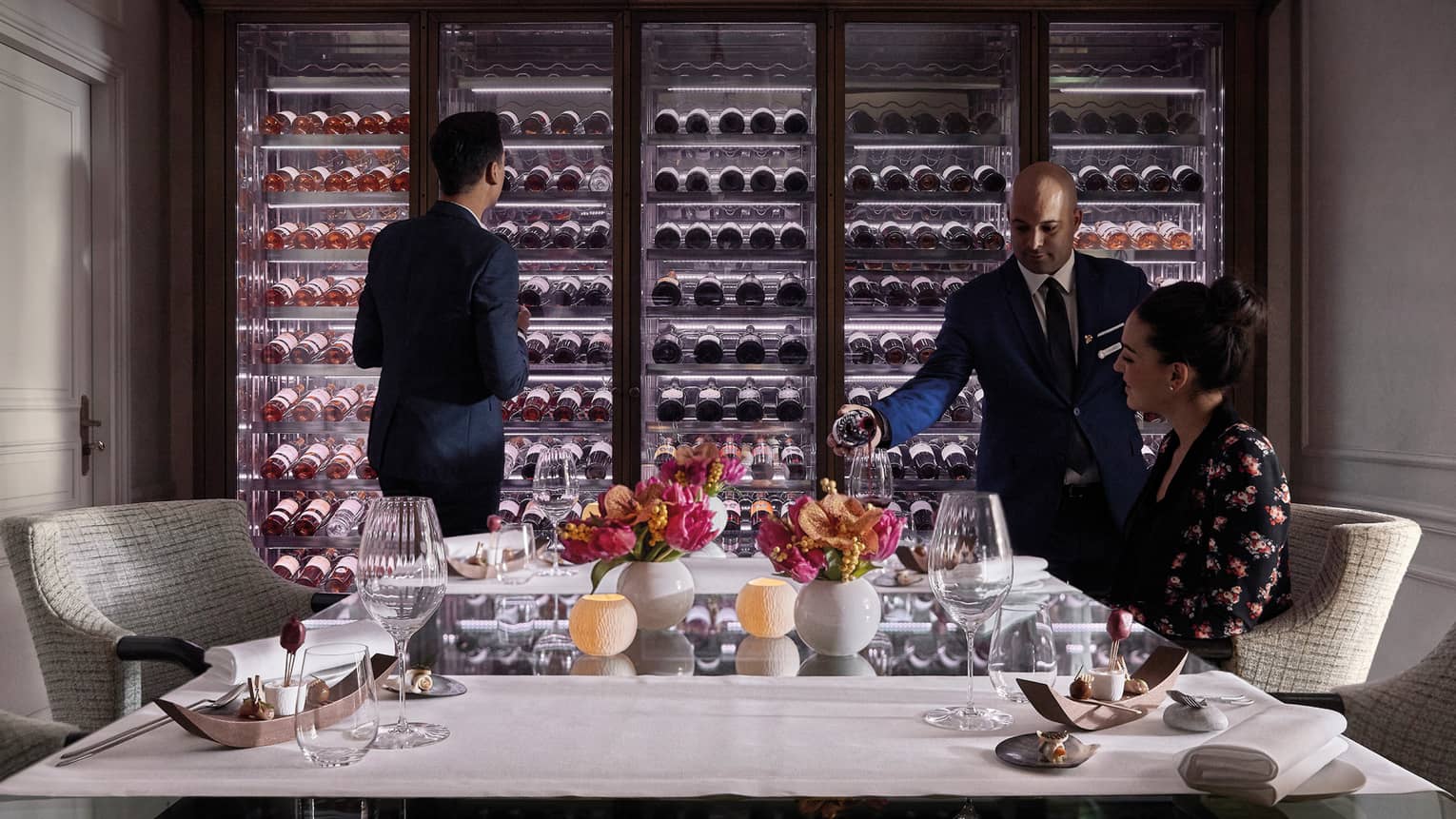 Man scans glass cellar as waitstaff pours wine in glass for woman at table