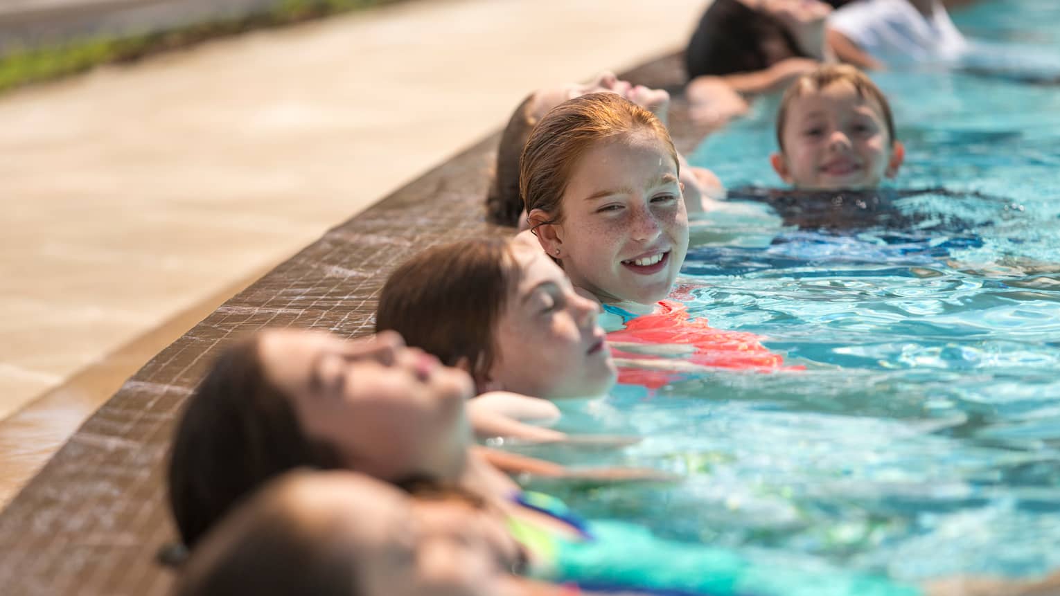 Six children smile, lean their heads against side of swimming pool