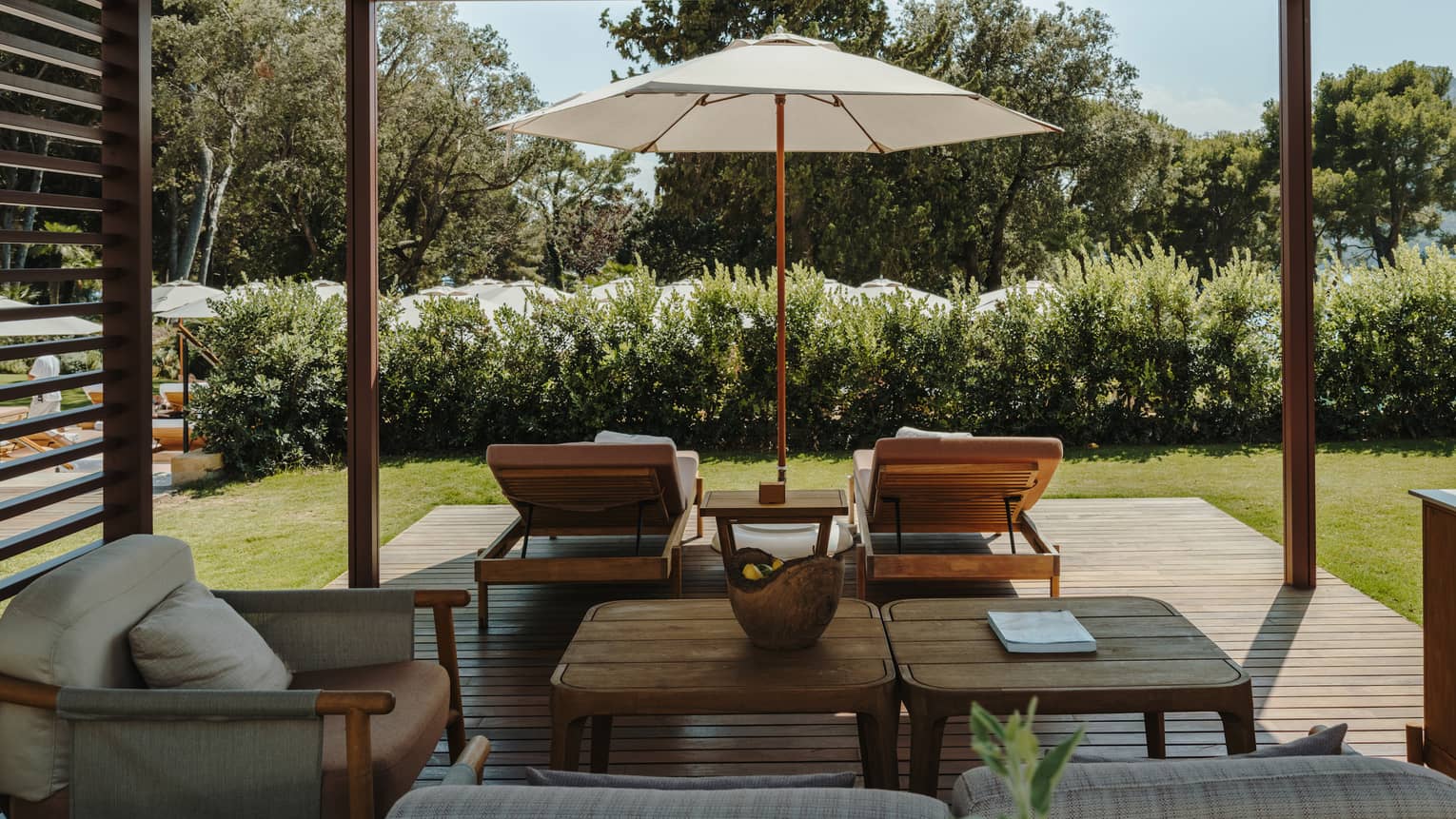 A shaded pool cabana with comfortable seating, a table, lounge chairs, an umbrella and lush green surroundings.