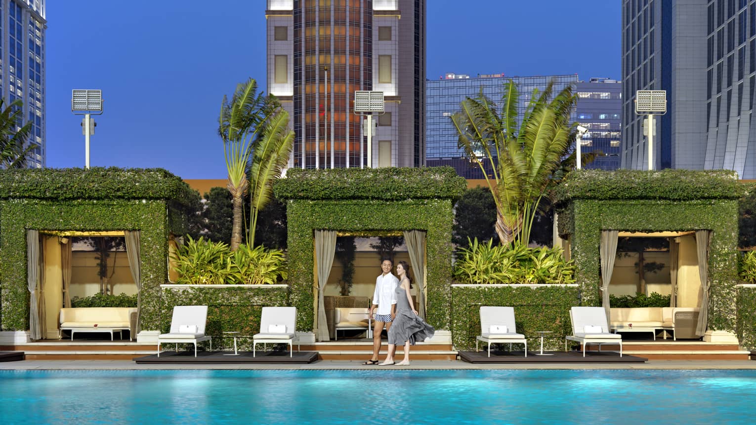 A couple smiles next to a pool, behind them a row of square archway cabanas covered in greenery and flanked by tall palms.