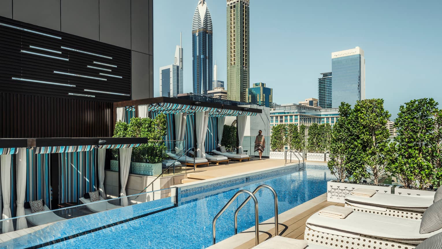 White lounge chairs, ladder into long blue swimming pool by cabanas on rooftop patio on sunny day
