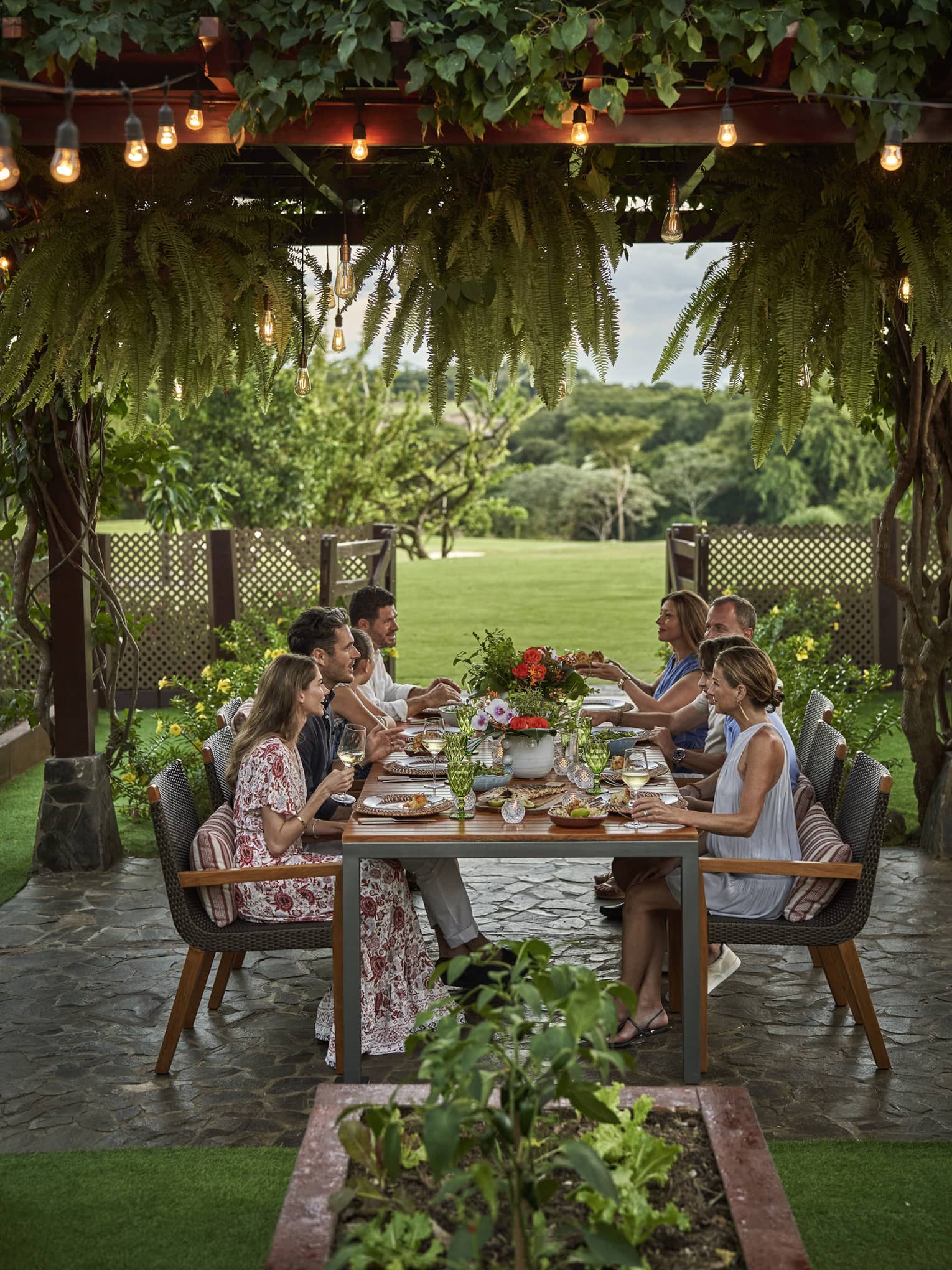 Eight people sit around a long dining table set up outside beneath a canopy of greenery and string lights