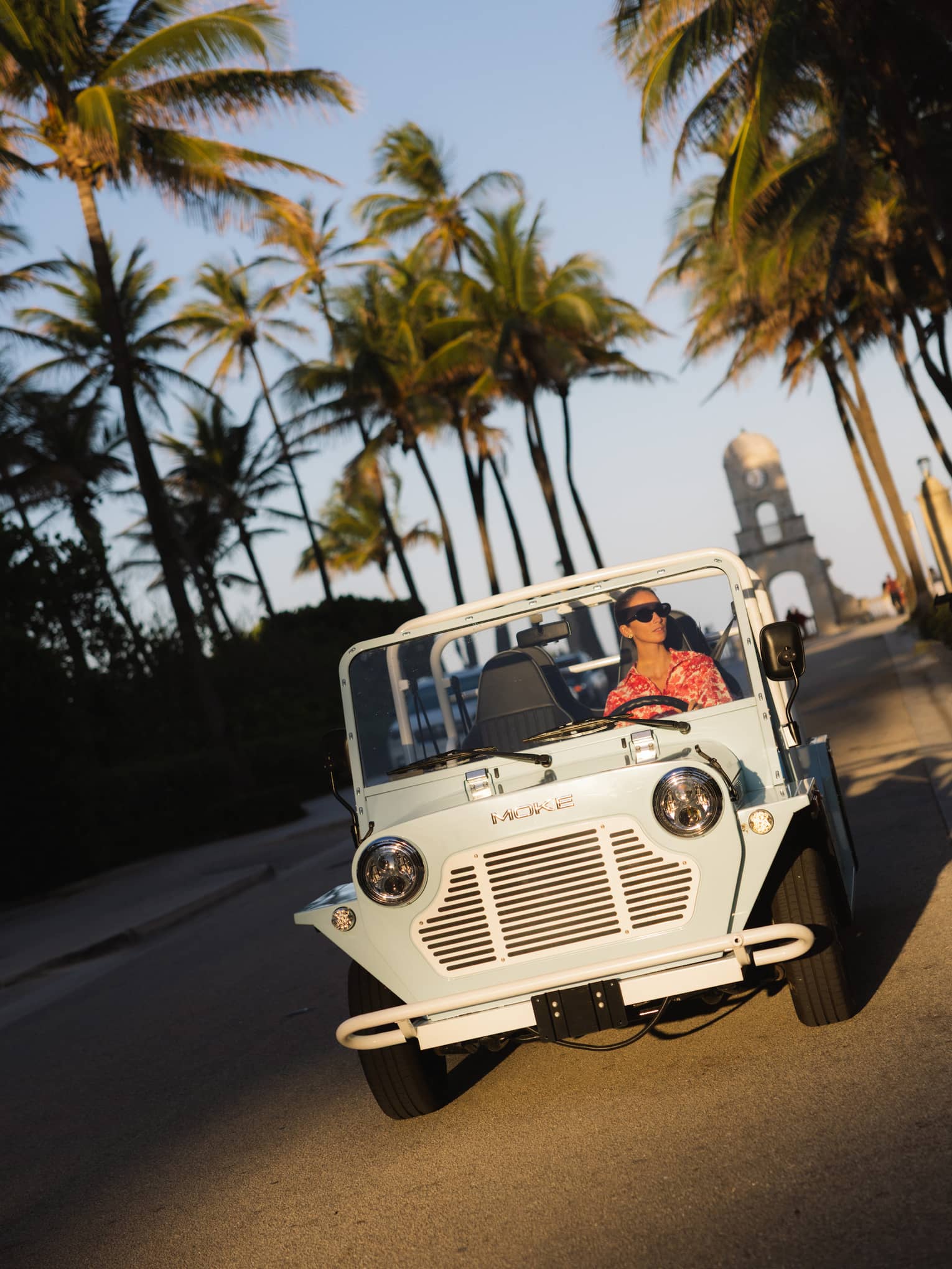 Guest wearing a red-and-white tropical shirt rides in a white MOKE along a palm tree?lined street at dusk
