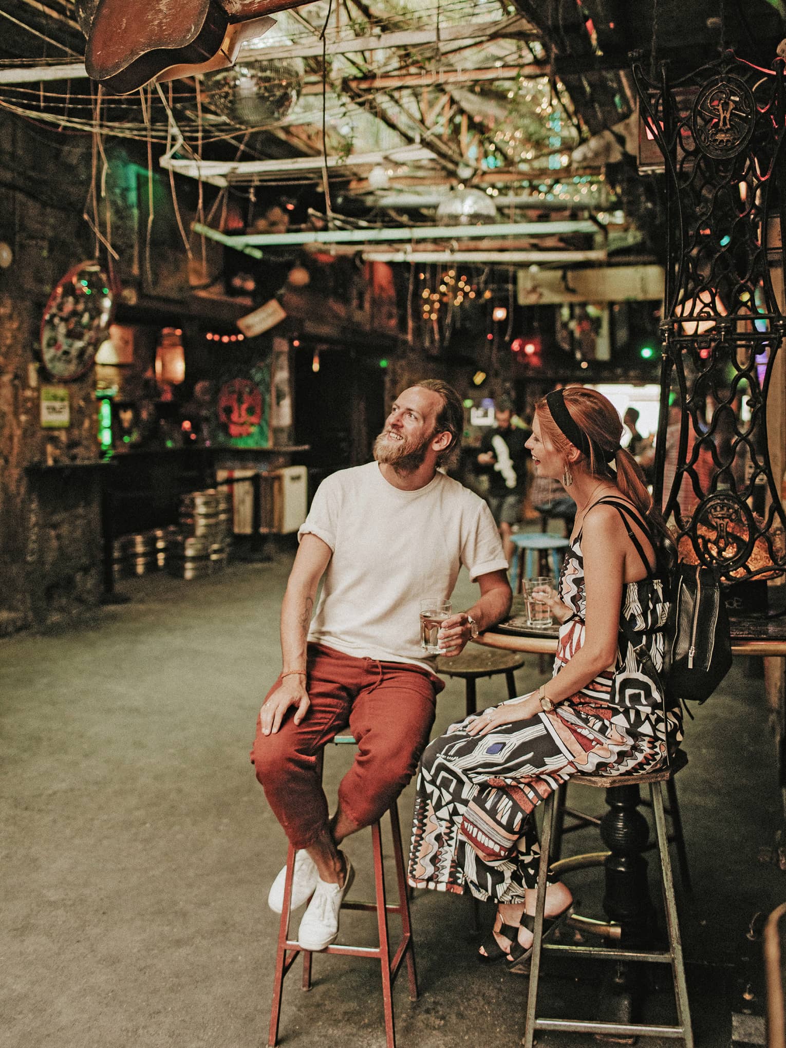 A smiling couple holding drinks, surrounded by myriad disparate objects and scrap parts on the walls and hanging overhead.