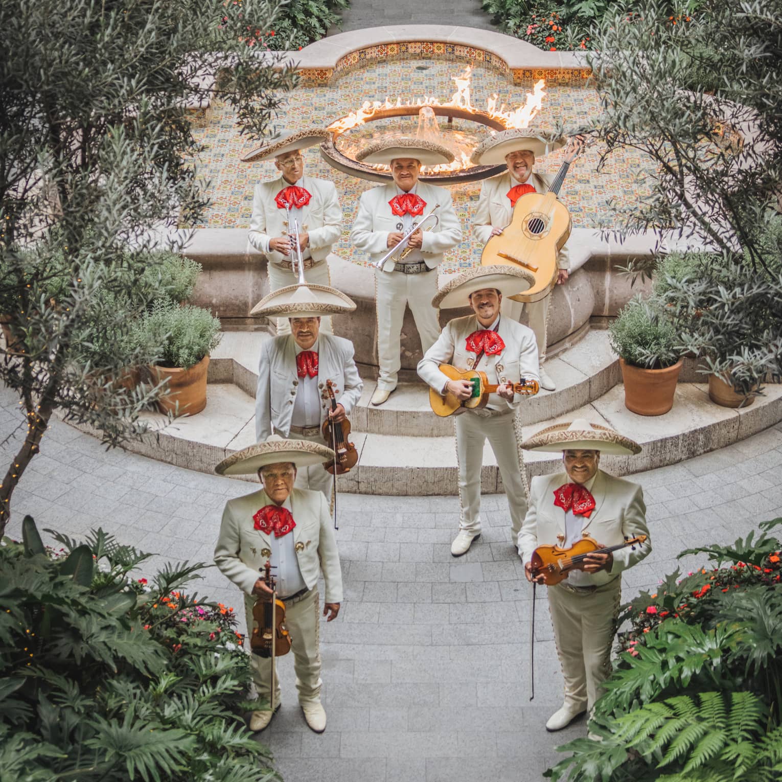 A group of mariachi musicians in an outdoor courtyard with paved pathways, abundant greenery and a fountain with a flame feature in the middle of it