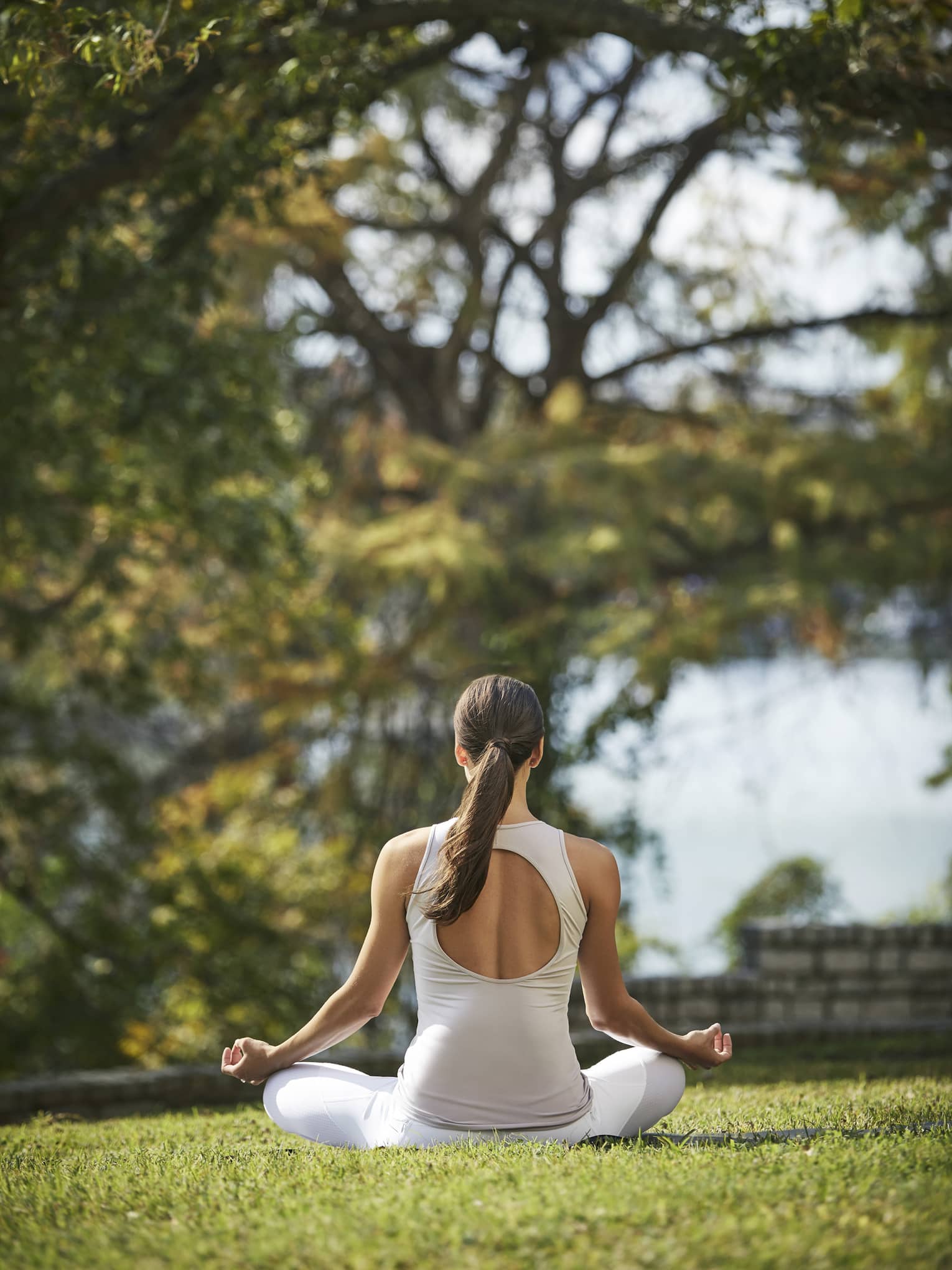 Rear view of a guest in white athletic wear meditating cross-legged with forearms rested on knees on a sunny patch of grass.