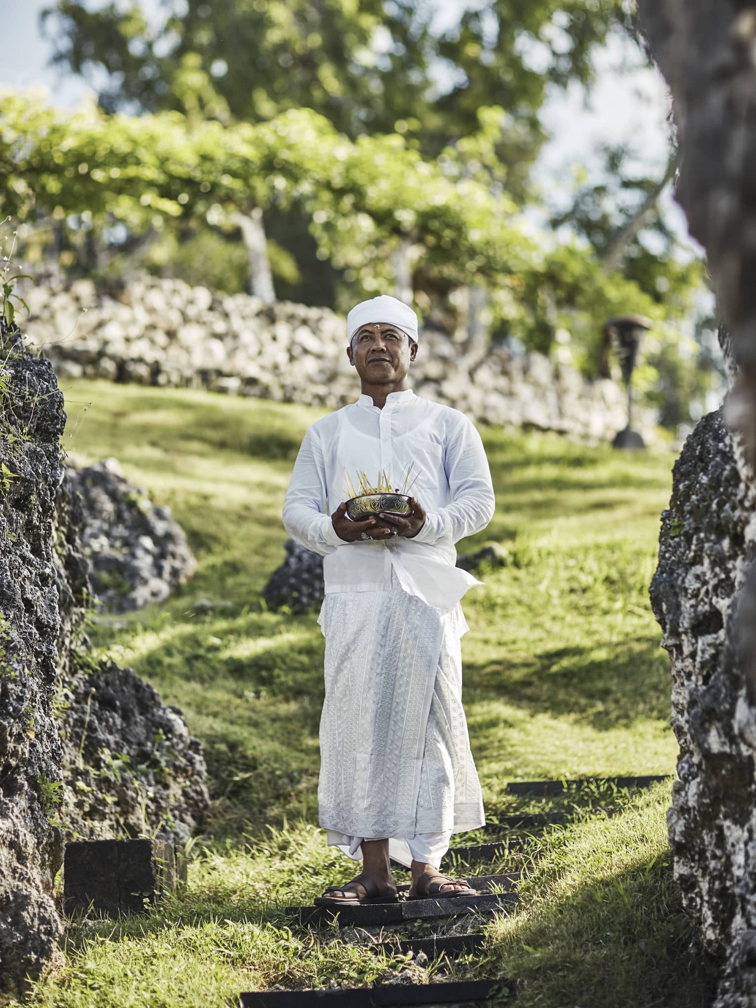 A priest in white attire stands on a stepping stone on a grassy hill, eyes gazing ahead, and holds a bowl of incense sticks.