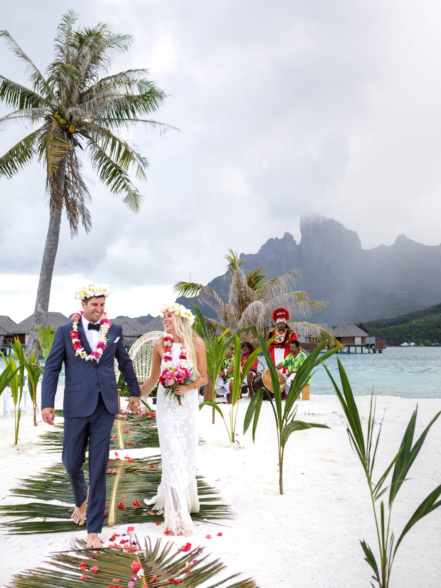 Bride and groom wearing floral crowns walk down aisle on white sand beach with palms