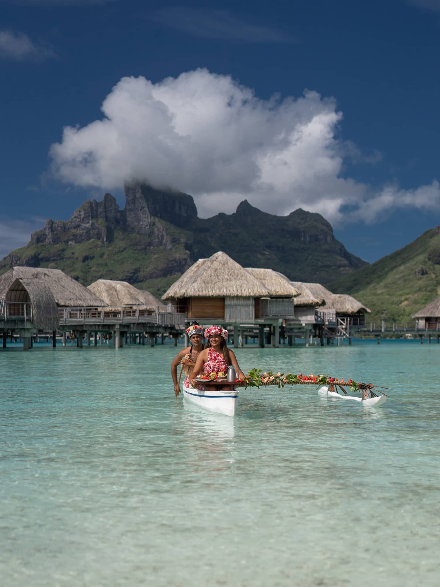 Two vibrantly dressed people in a canoe proffer a tray with colourful food attached to a pole draped with leaves and flowers.