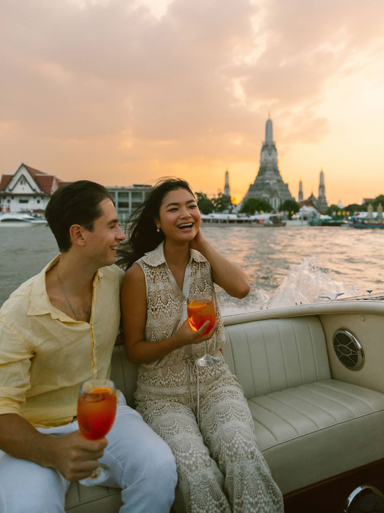 Smiling couple sits at back of boat holding cocktails on Chao Phraya River sunset tour