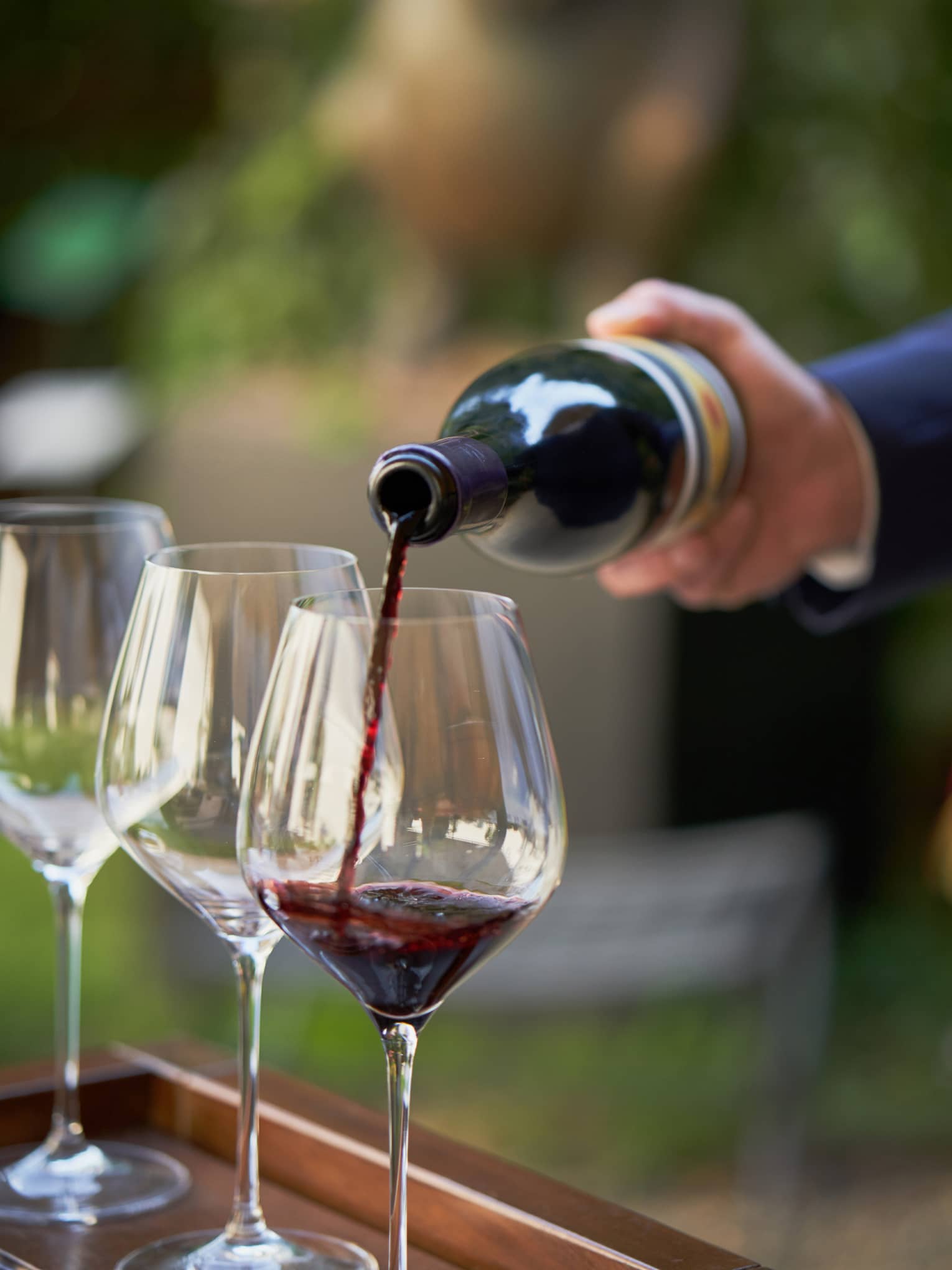 A close up of hands pouring a bottle of red wine into a clear wine glass that rests on a wooden tray, outdoors
