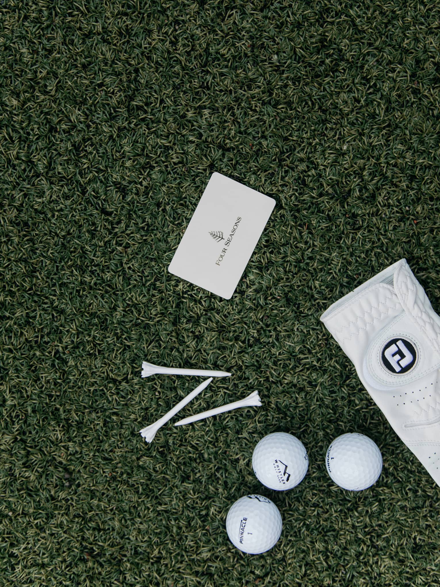 On green turf, a Four Seasons card, three golf balls, three tees and a white leather golf glove dotted with tiny holes.