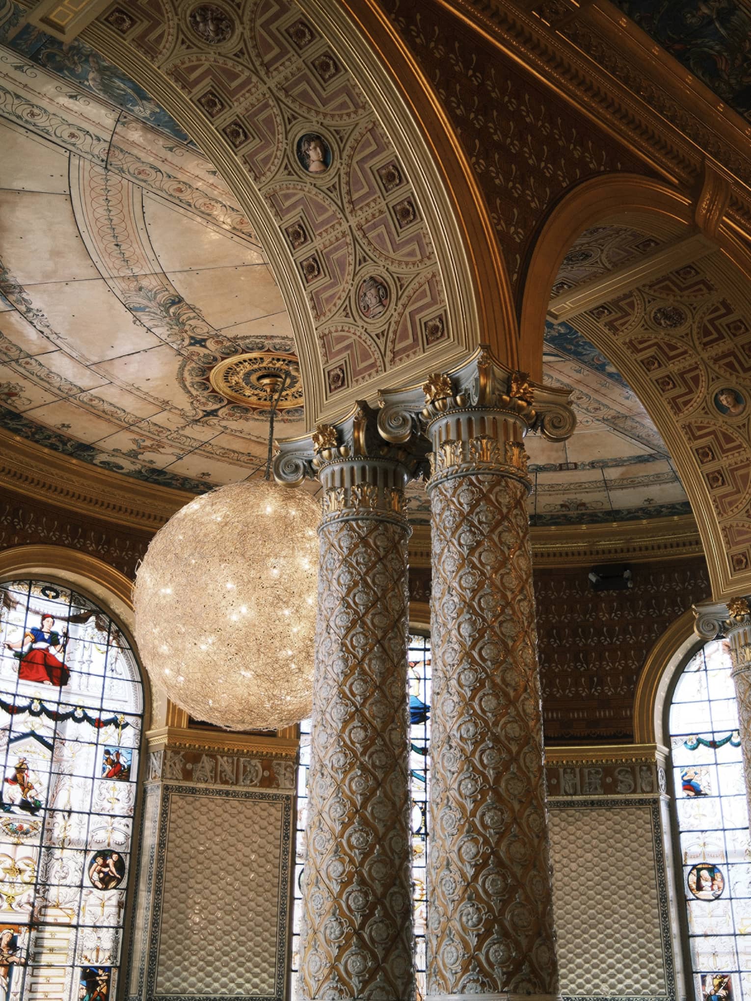 An interior space with a high arches and columns with decorative inlay and carving. Stained-glass windows, decorative ceiling and walls, and chandelier lighting are in the background.