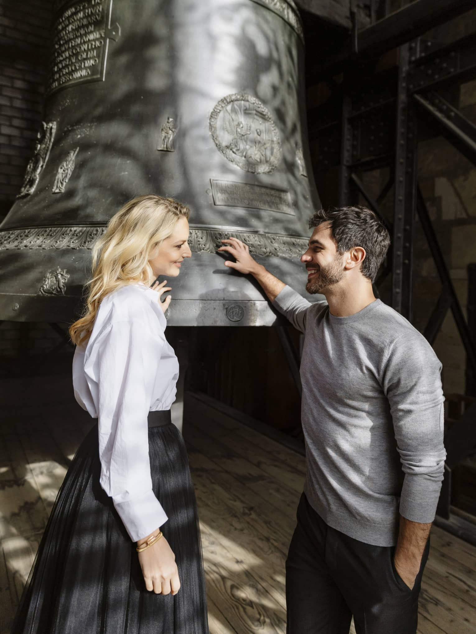 Two people stand next to a large metal bell in the bell tower of a cathedral.
