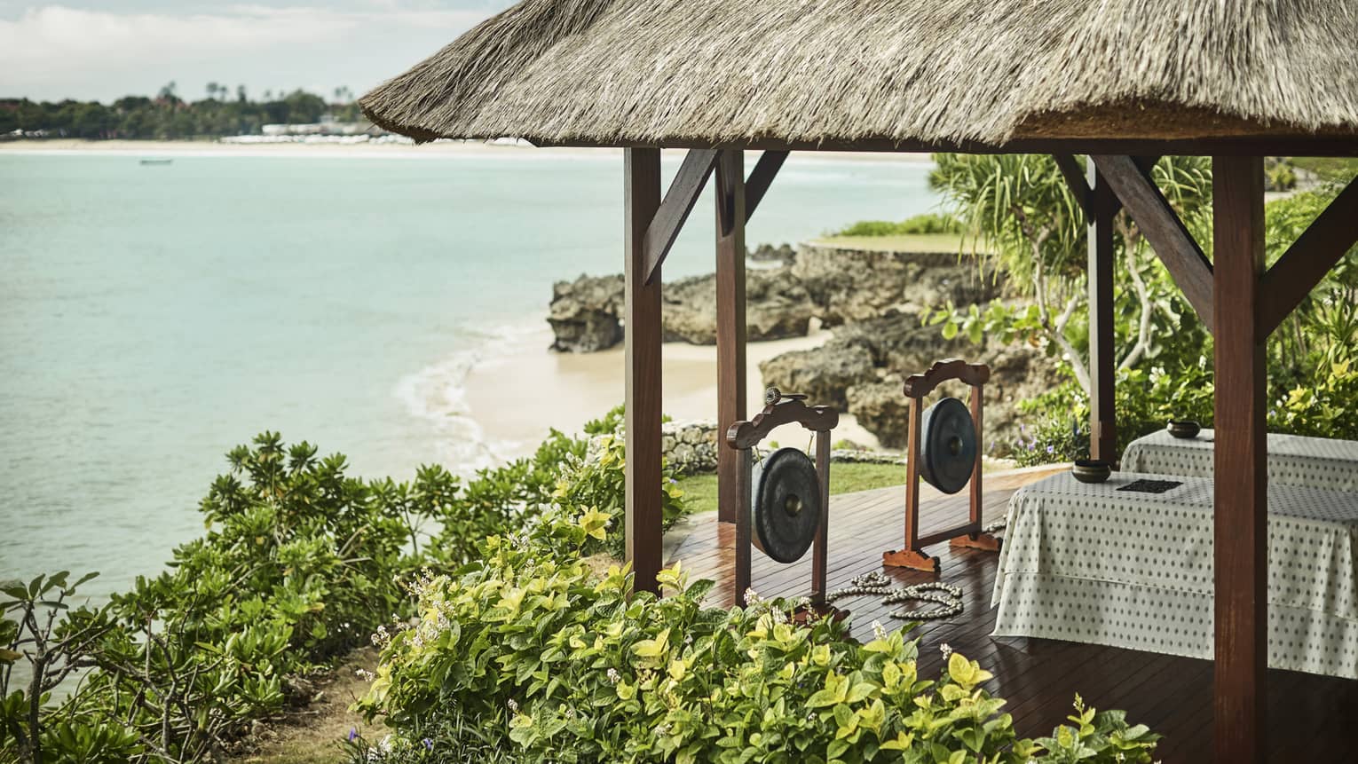 A thatched hut with two tables and two gongs overlooks calm, teal water and a beach lined with rocks and lush greenery.