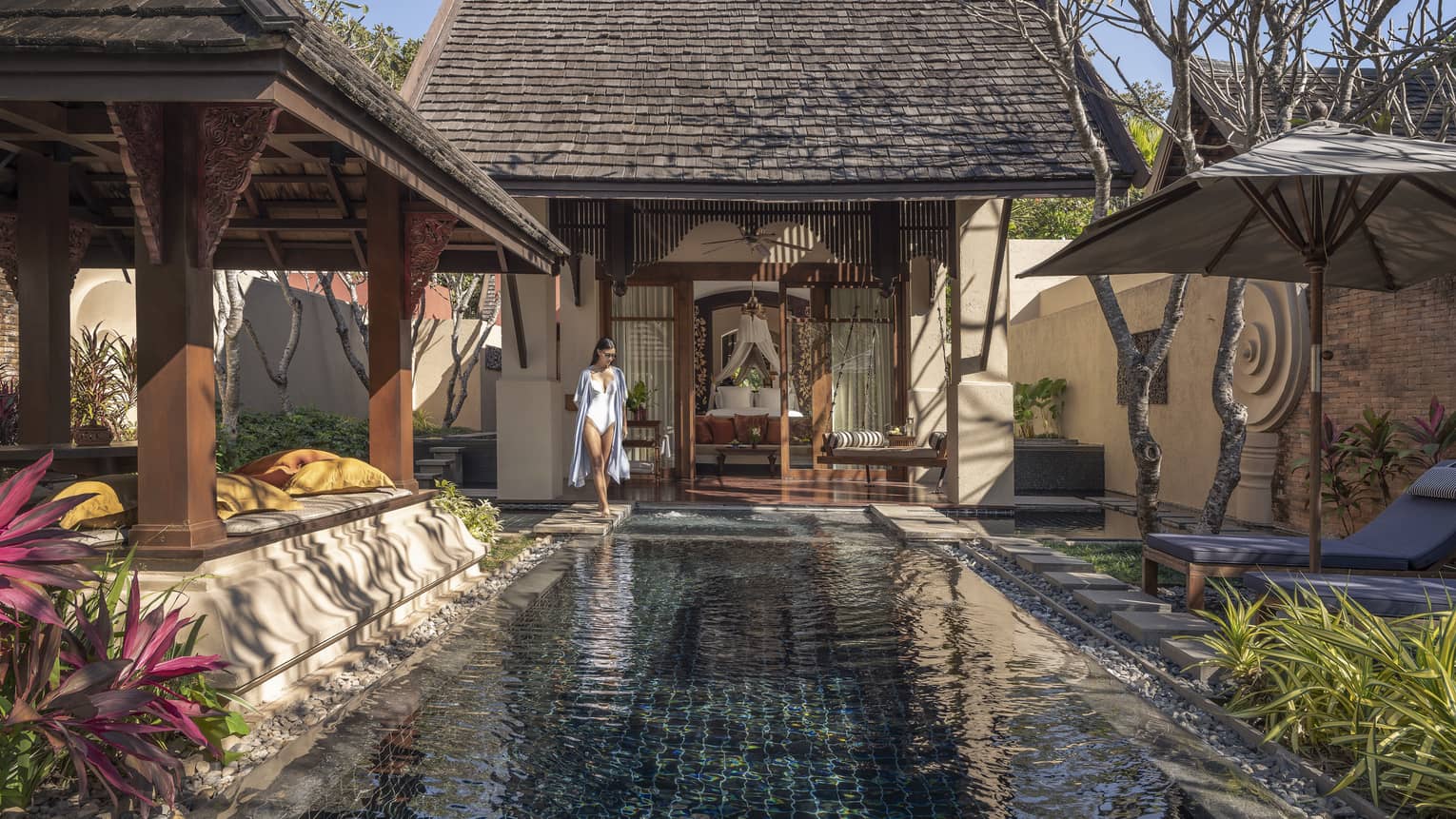 Person walking beside a private pool in a courtyard with sun loungers, tropical plants and traditional wooden architecture