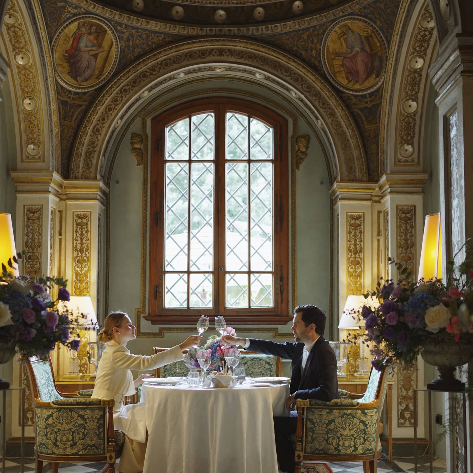 Two people seated at an elegant dining table in an ornately decorated room with high ceilings, patterned walls, and a large arched window, toasting with glasses.