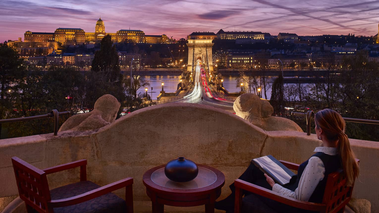 Woman sits on balcony of Royal Suite overlooking bridge and river at dusk 
