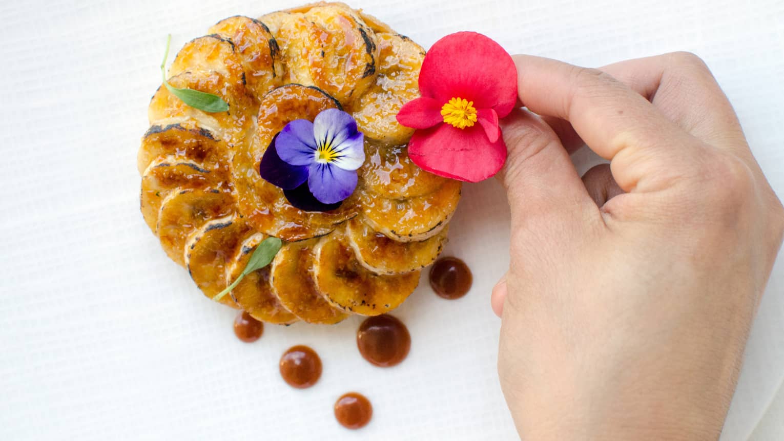 Close-up of hand placing flower garnish on ring of glazed plantain banana slices