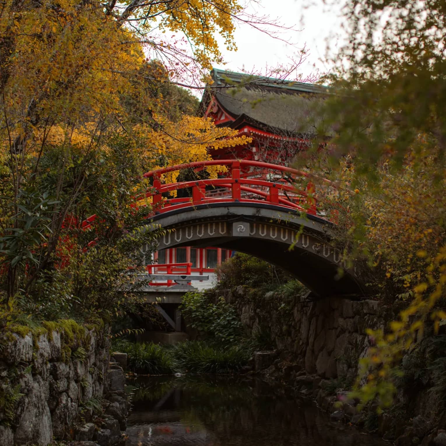 Arched bridge over a creek and temple surrounded by autumn foliage