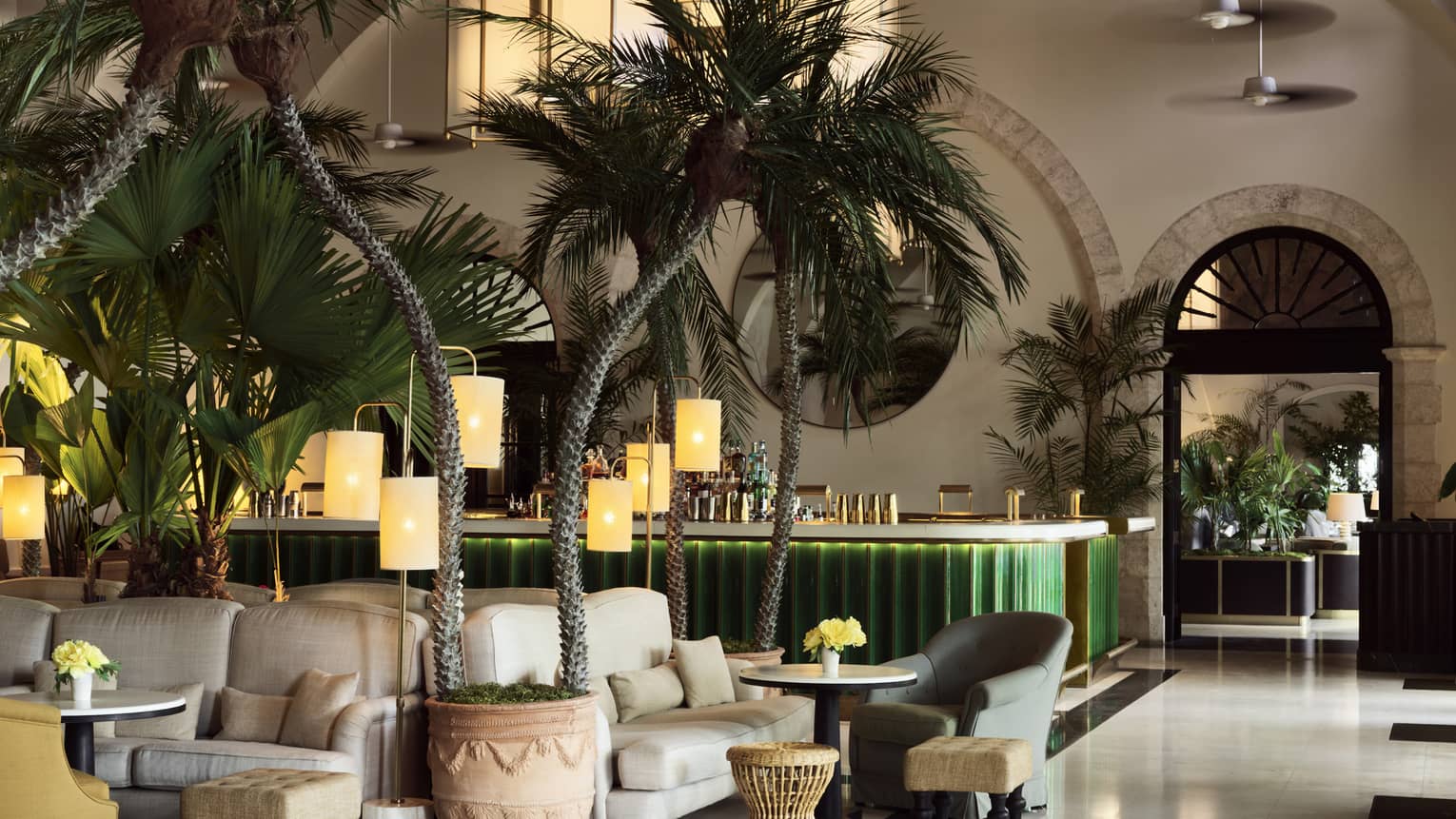 Indoor lounge with palm trees, white upholstered arm chairs, green bar in the background