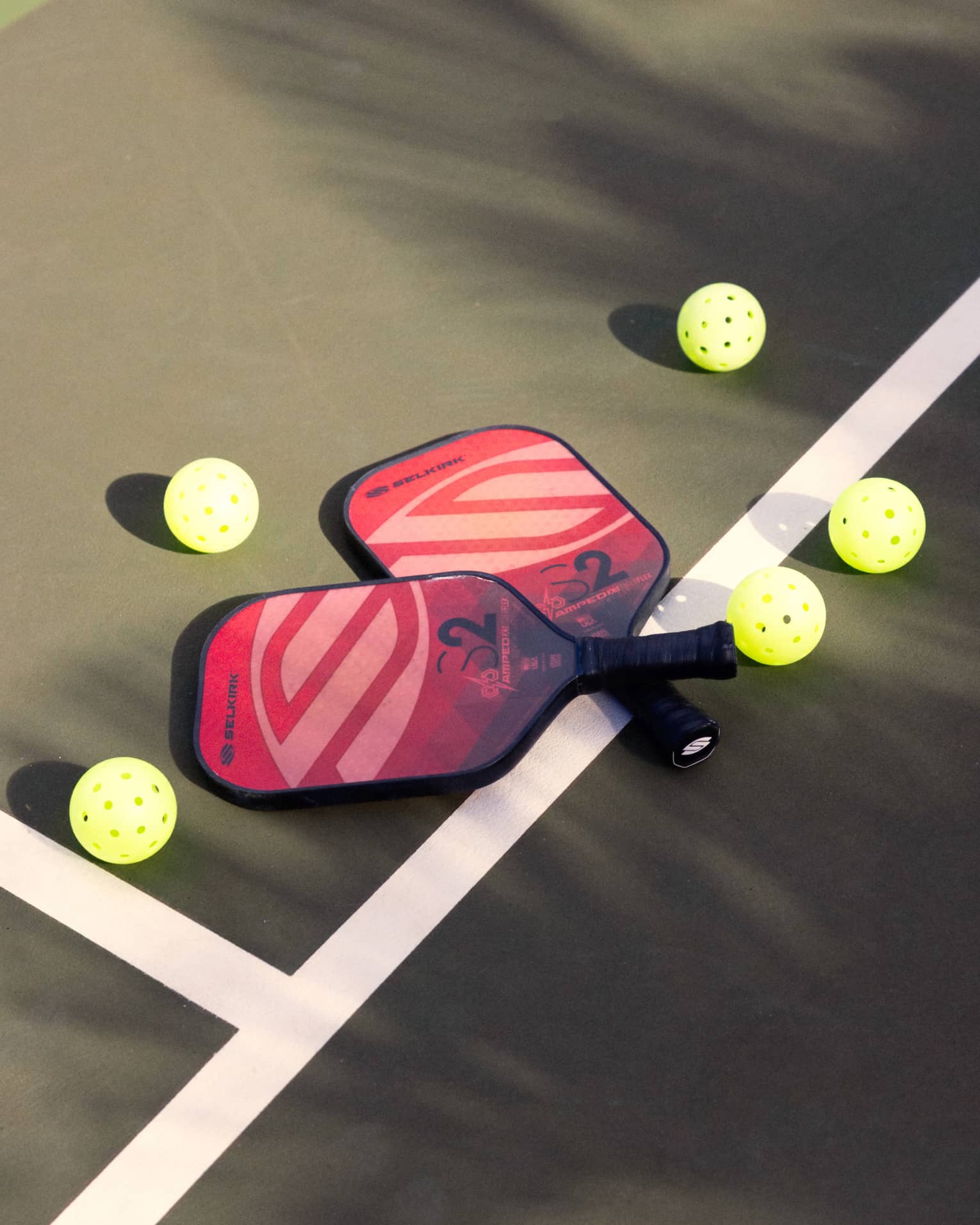 Close-up of two red and pink pickleball racquets, handles crisscrossed on a white court line, surrounded by five pickleballs.