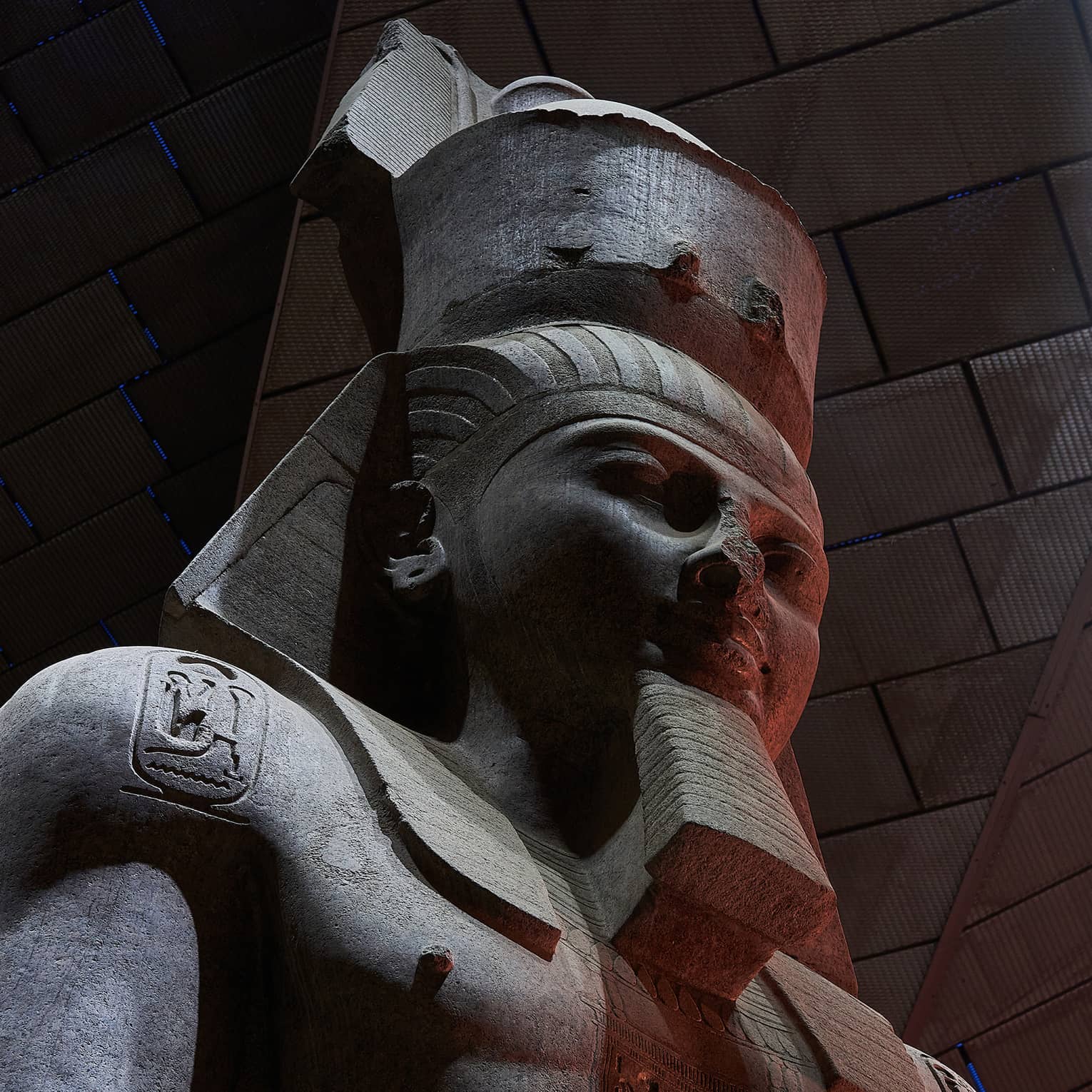 Close-up on a dark stone statue of a bearded pharoah with three heiroglyphic carvings on his chest and shoulders.