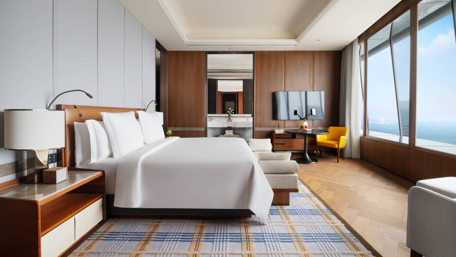 Large hotel room suite with king bed, plaid-patterned rug, full wall of windows with ocean view, and arm chairs
