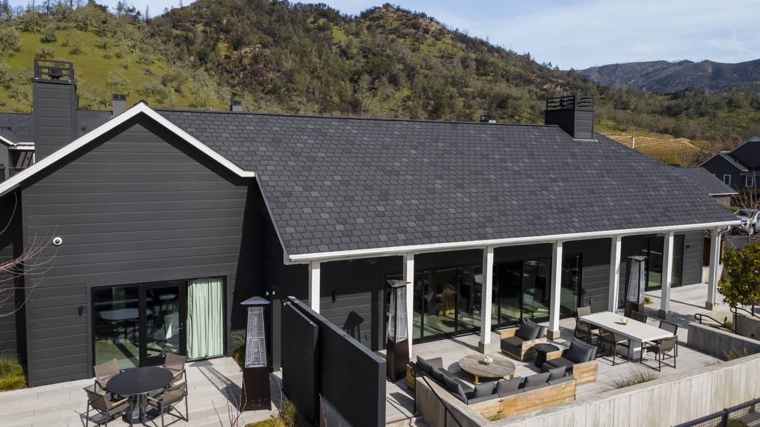 Aerial view of a luxury resort villa with dark siding, a large patio featuring outdoor seating areas and dining tables, set against a backdrop of green hills