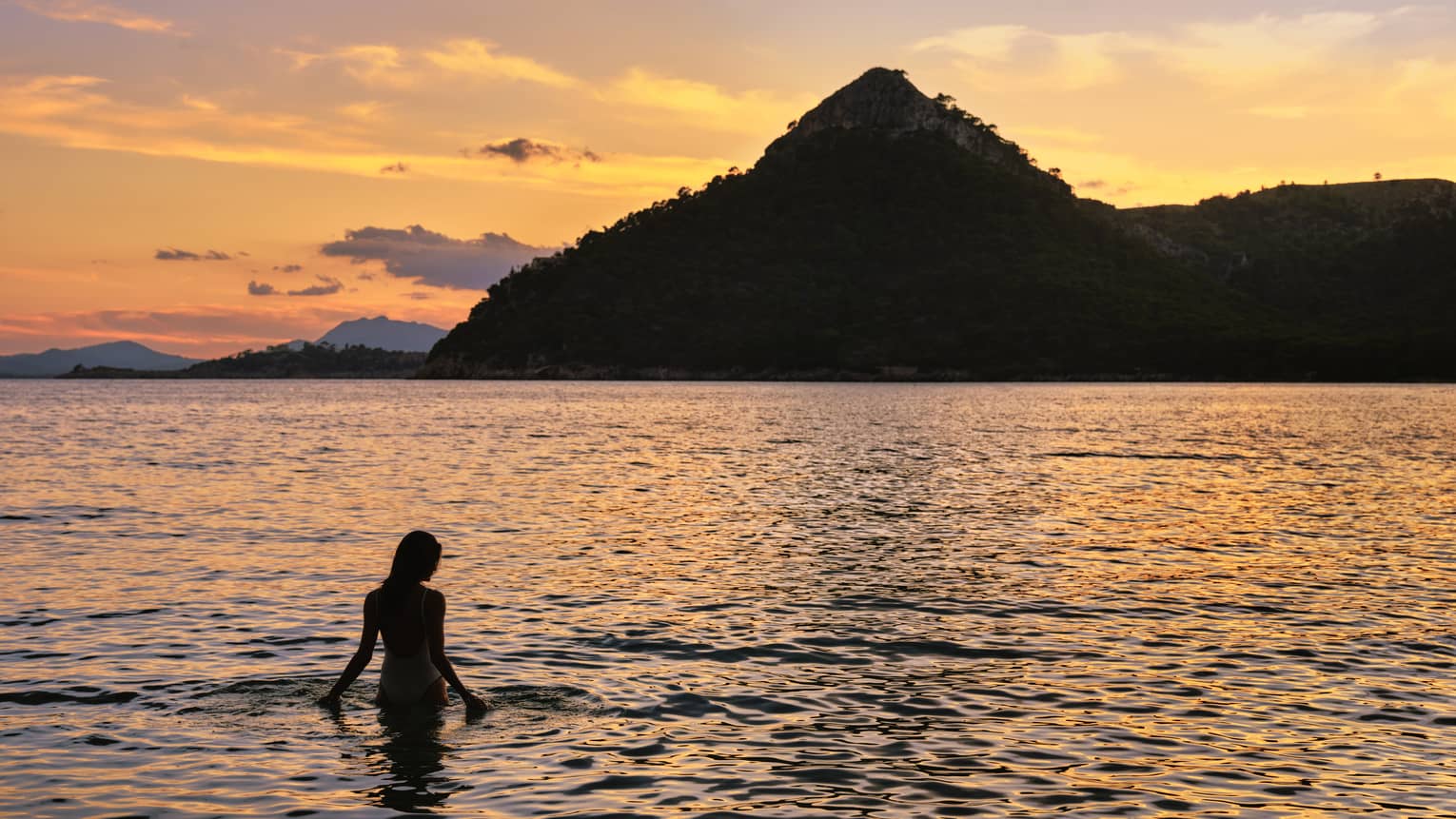 A guest silhouetted in a fiery sunset wades into rippling water against the dramatic backdrop of a towering hill.