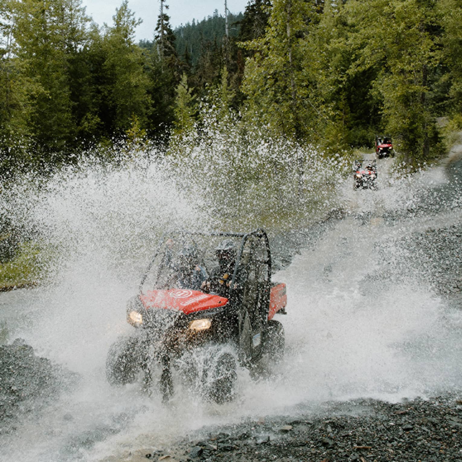 A red-and-black ATV driving through water, creating a thick veil of water spray against a background of pine forest.