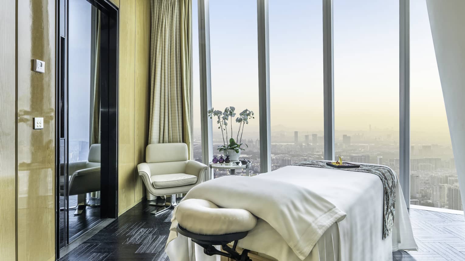 A bright massage room with opulent bedding and a side table with white orchids and glass vials, overlooking a hazy cityscape.