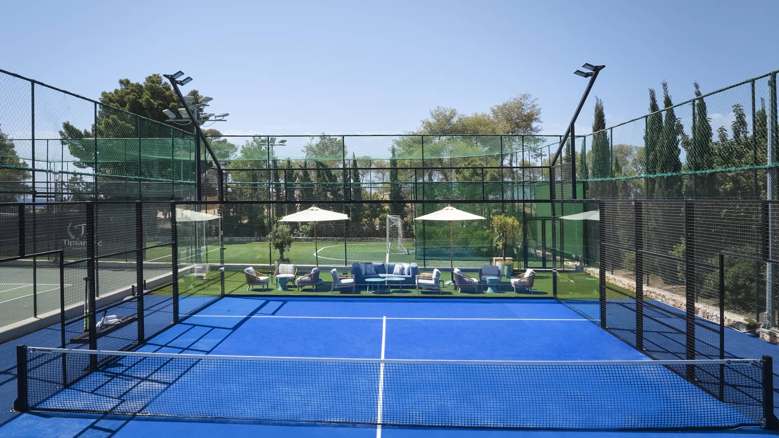 Bright blue padel court surrounded by fencing, white umbrellas and lounge chairs