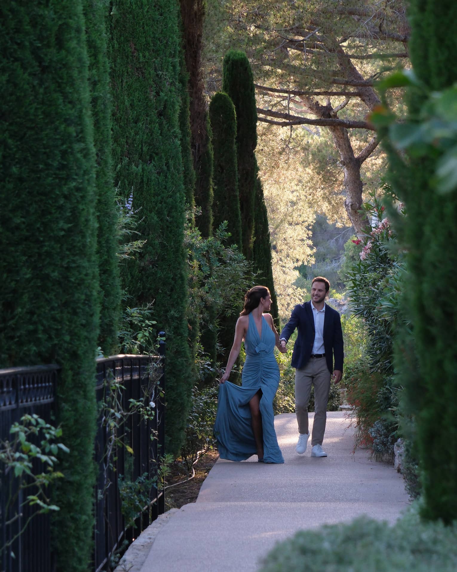 A couple dressed in formal attire walks along a shaded pathway surrounded by tall, lush trees