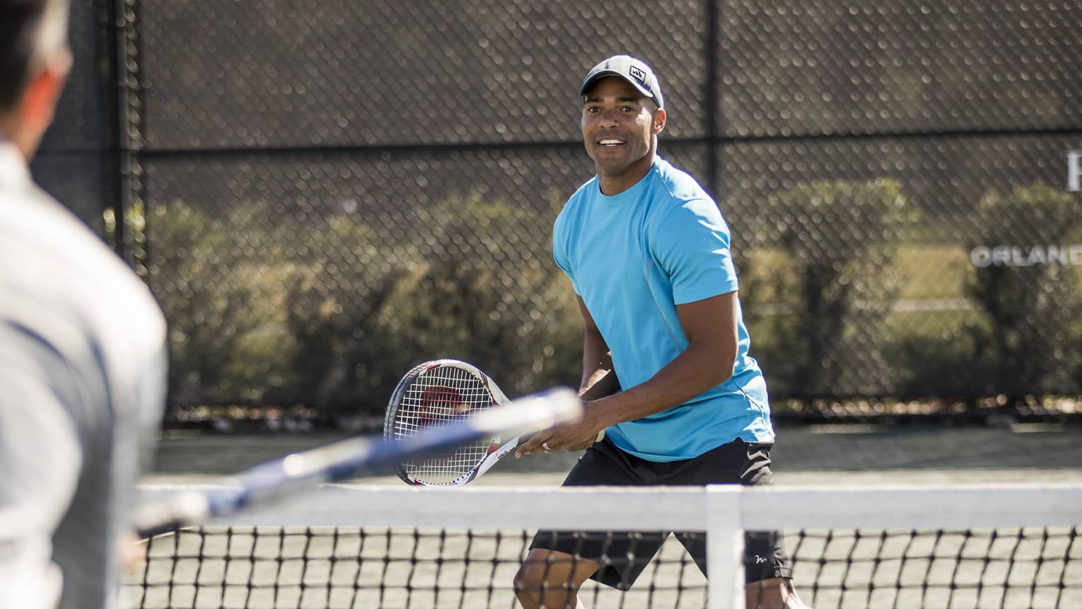 Man wearing blue T-shirt and hat holds racket as tennis ball flies over net