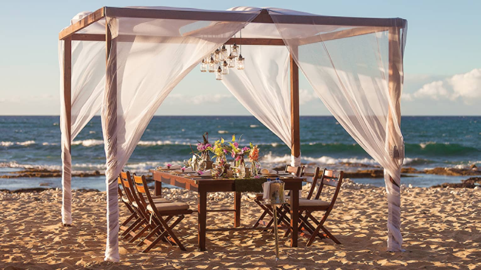 Wood dining table with fresh flowers under sheer white canopy, lanterns on beach