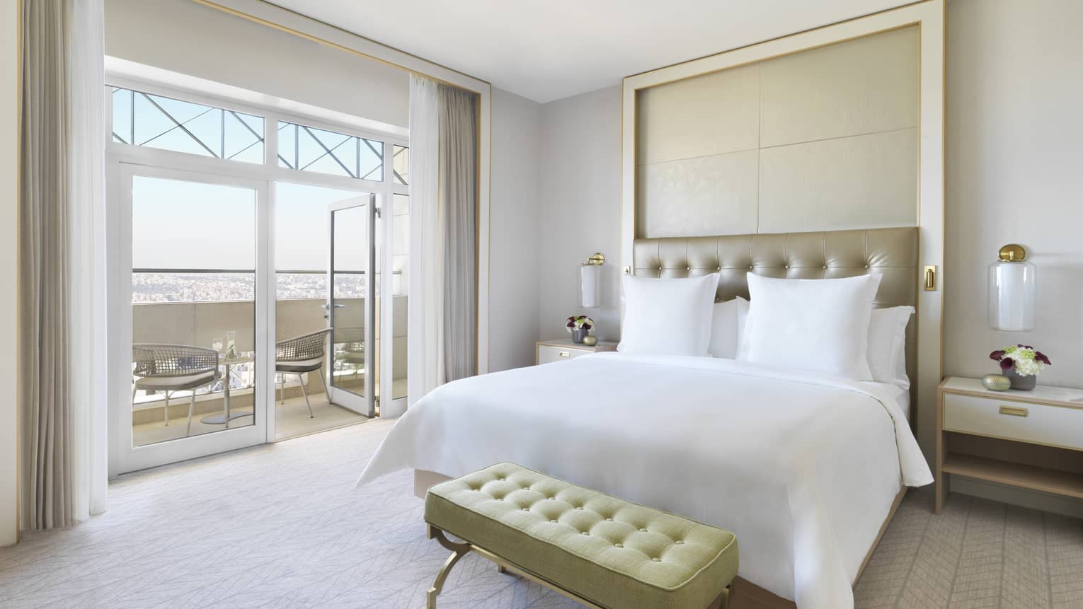 A plush white bed in the centre of the room with a yellow ottoman at the foot. The room opens up to a veranda overlooking the city. 