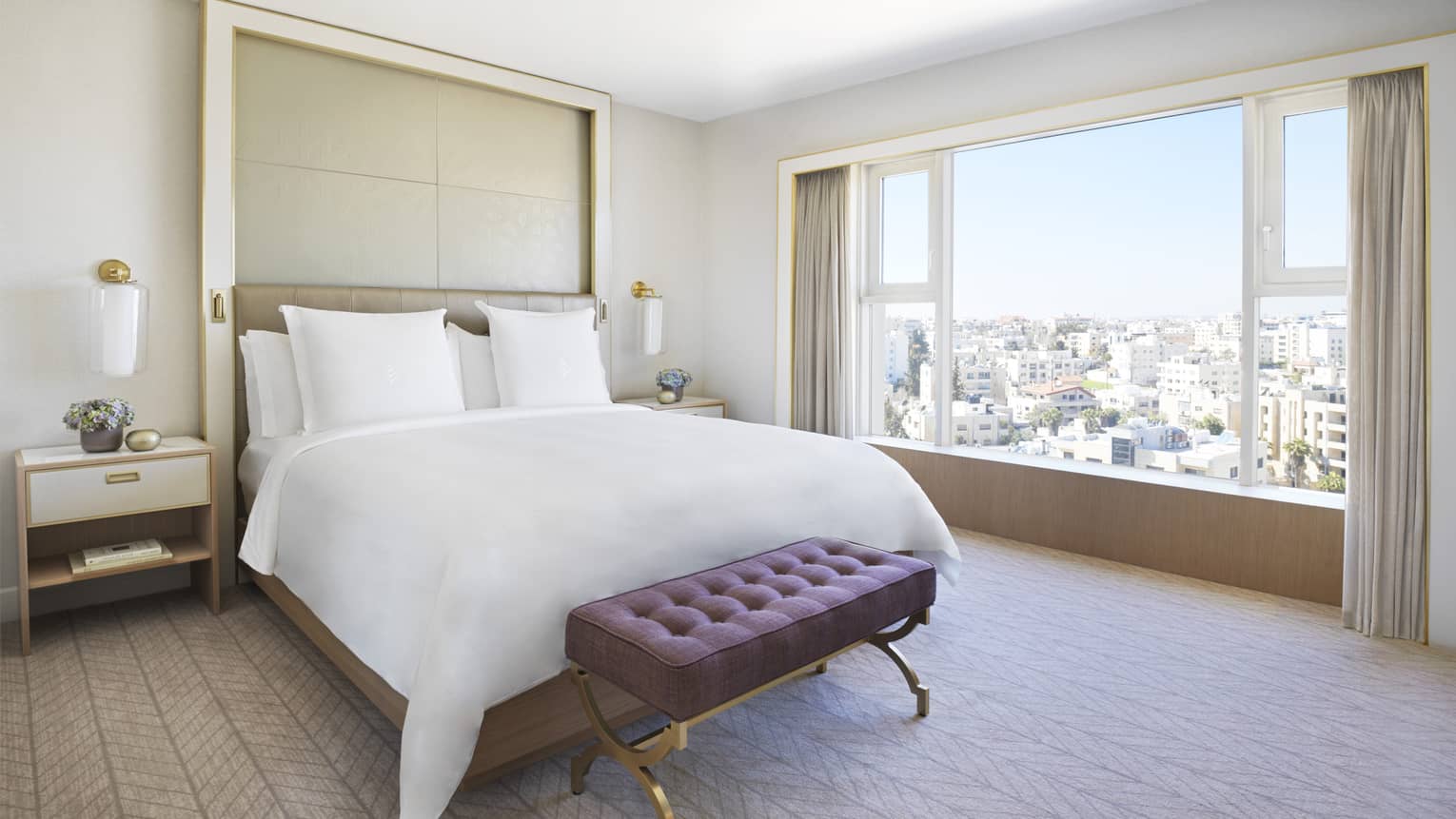 A white bed in the centre of the room with a purple ottoman at the foot. The room overlooks the city. 