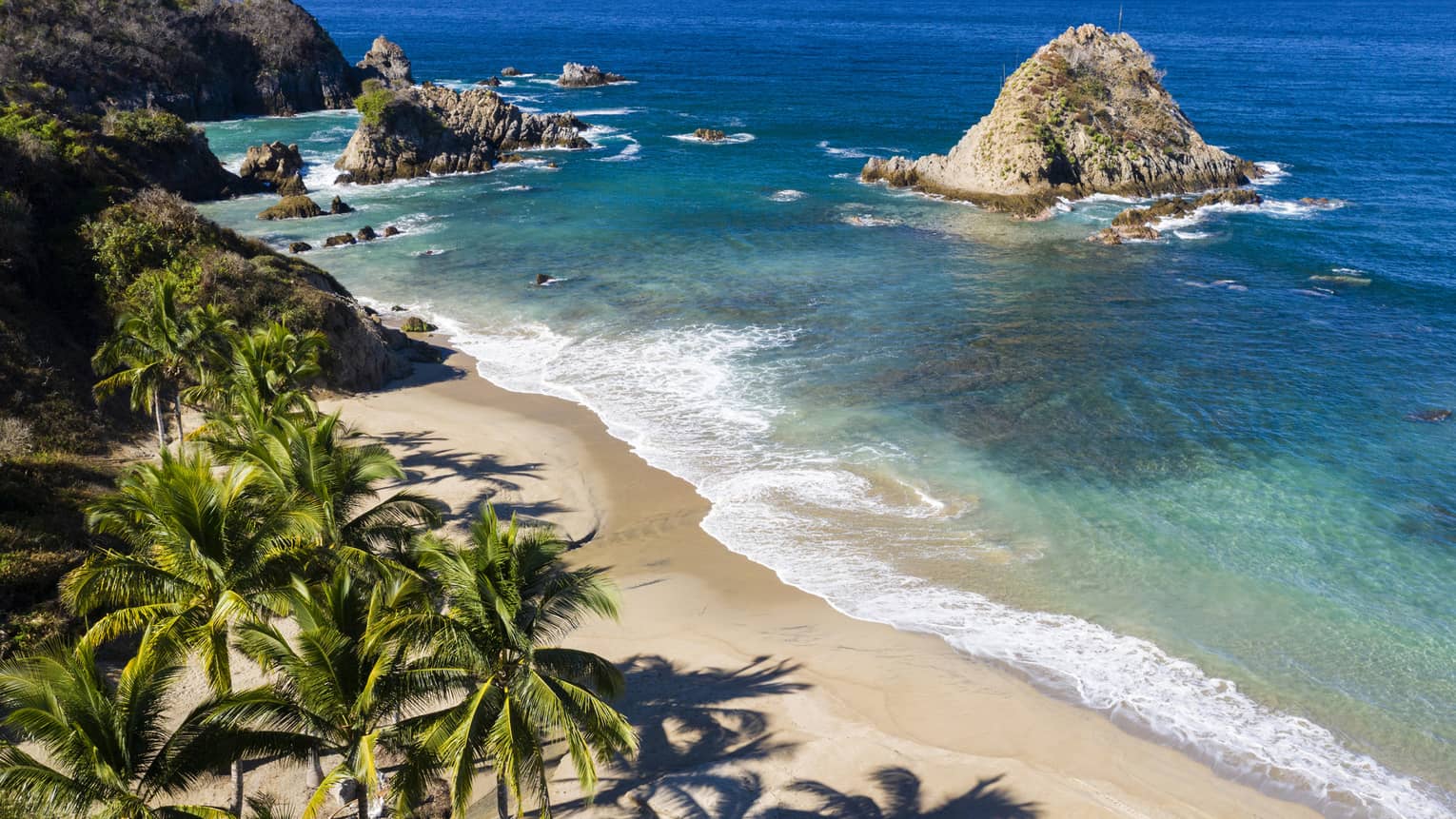 Aerial view of beach with craggy shore, palm trees, two-tone blue water