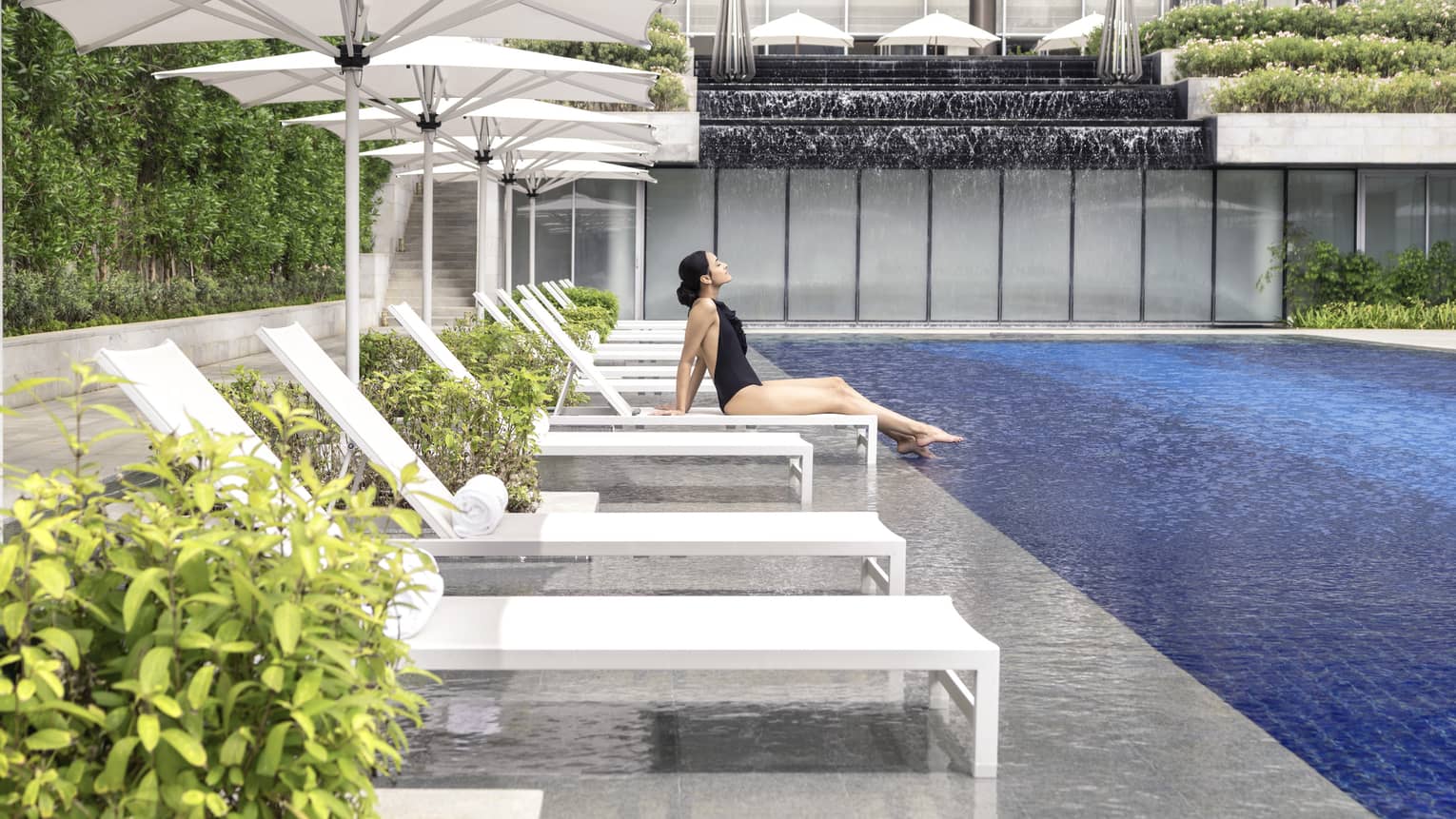 A woman in a black bathing suit relaxes on a chaise by the pool