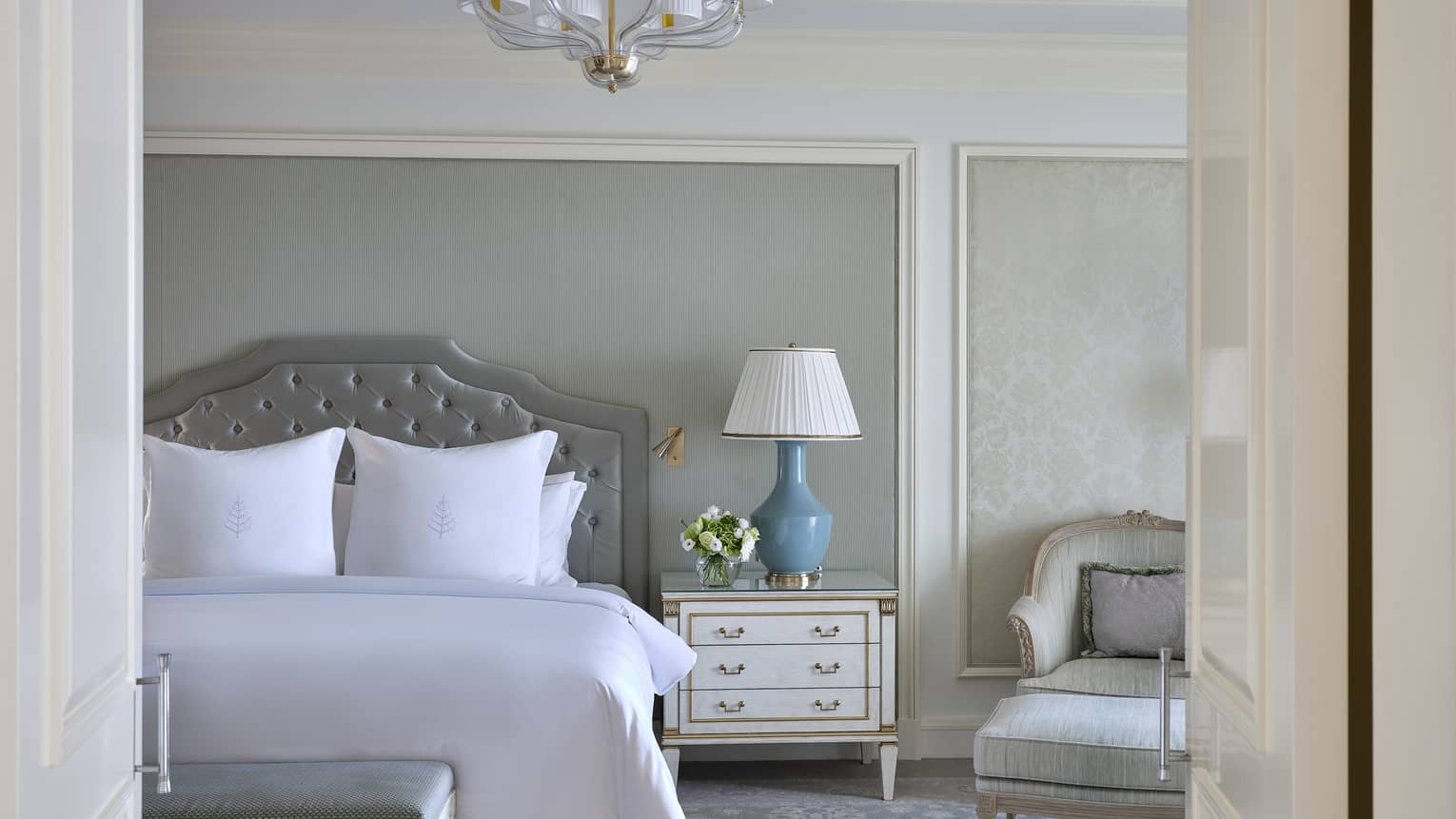 Elegant hotel room featuring king bed with white sheets, chandelier, grey headboard and crown molding
