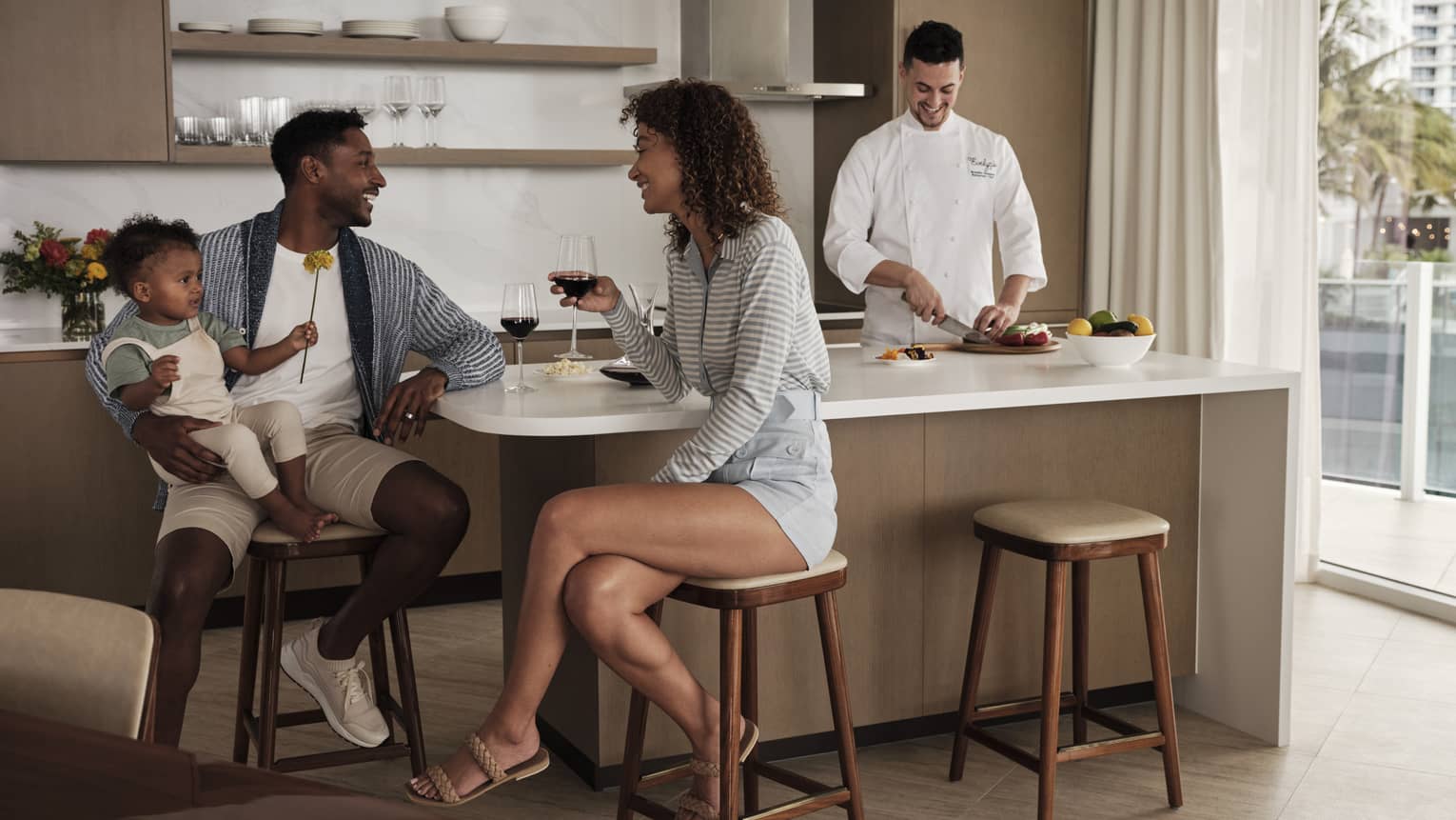 Parents sit at a kitchen island, toddler in dad's arms, laughing over glasses of red wine while a chef prepares a meal. 