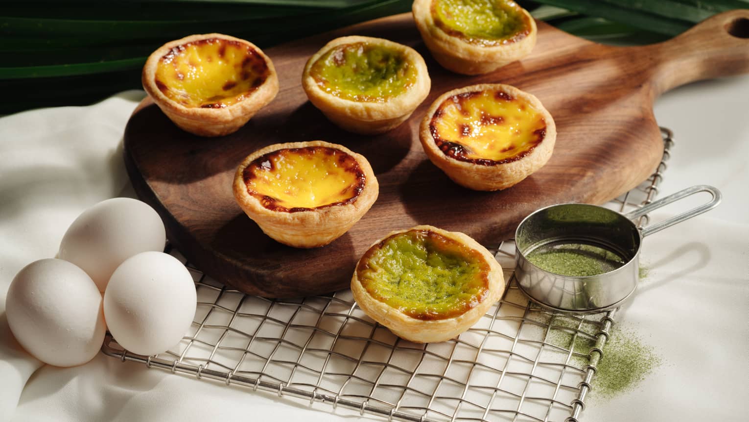 Six custard tarts on a wooden board over a wire rack. Four have golden brown surfaces, and two are green. Next to the board are three white eggs.