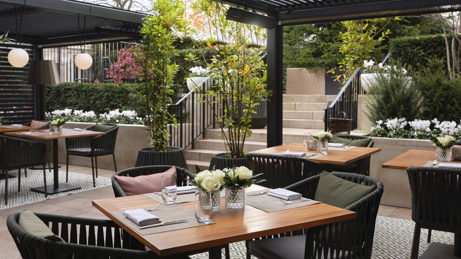 Set square wooden tables, dark rattan chairs, outdoor diamond-patterned rug under pergola