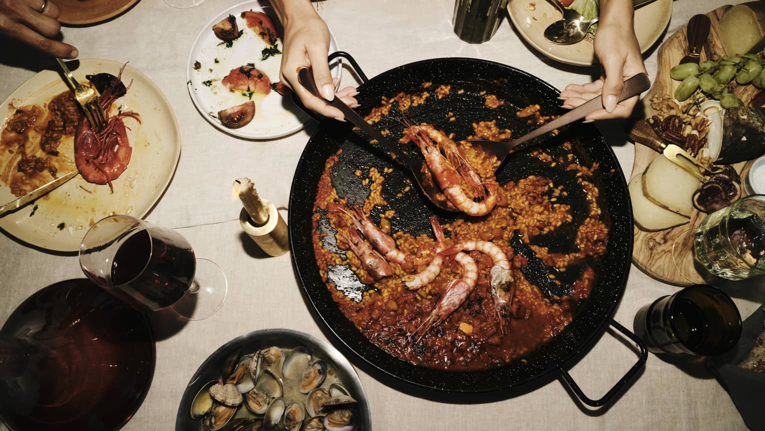Two hands use large spoons to lift prawns from a saucy paella dish set amid plates of seafood, cheese and fruit.