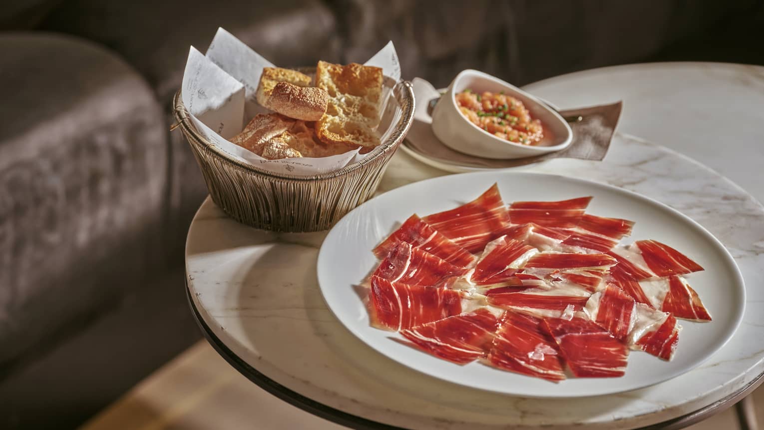 Plate of Iberico ham slices beside bowl of crackers on small marble-top table