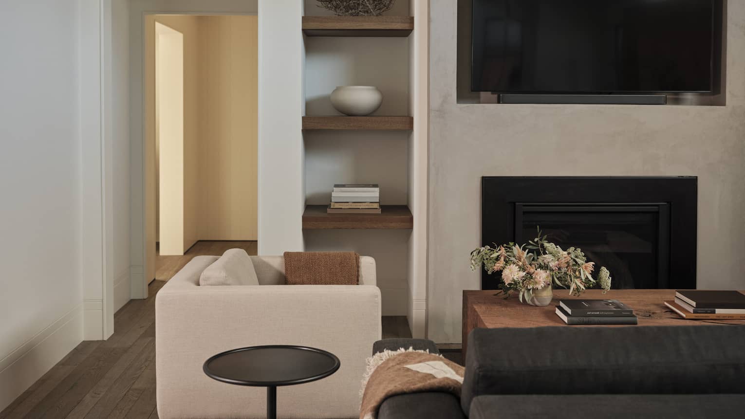 Living room with gray sofa, beige armchair, side table, wooden coffee table with floral arrangement, built-in shelves with decor, wall-mounted TV and fireplace