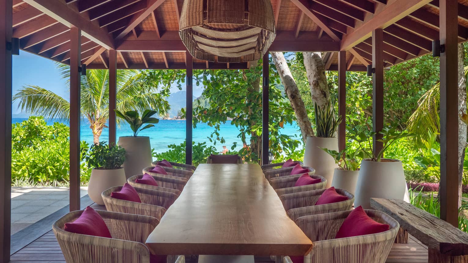 Natural wood dining table with 10 chairs underneath an outdoor pavilion with sea view