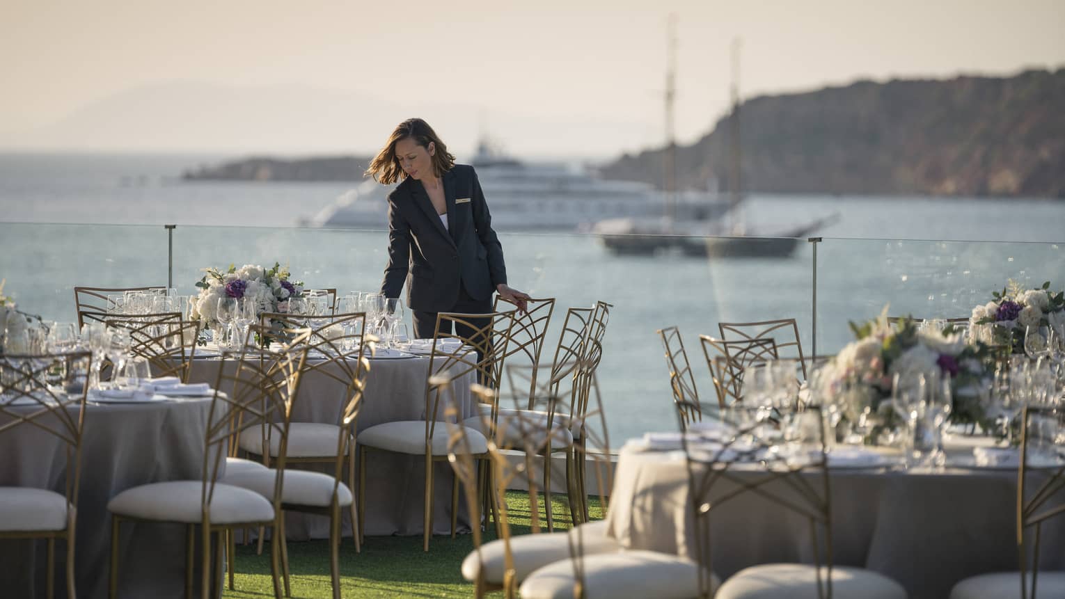 Woman arranges round table with glasses, flowers, white and gold chairs overlooking ocean and boat