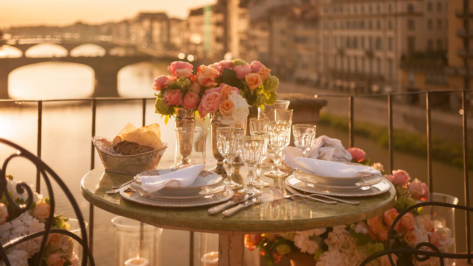 A romantic table for two is set with white porcelain plates, crystal glasses, pink roses on a balcony overlooking a bridge and historic building in Florence